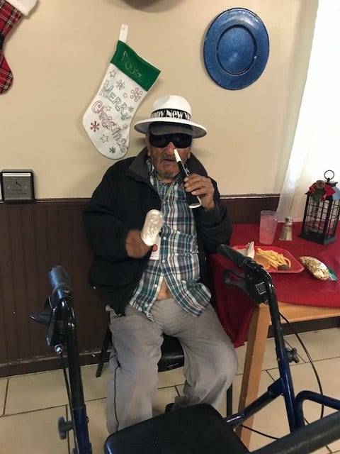 A man wearing a hat and sunglasses enjoys a drink while sitting in a wheelchair, surrounded by holiday decorations.