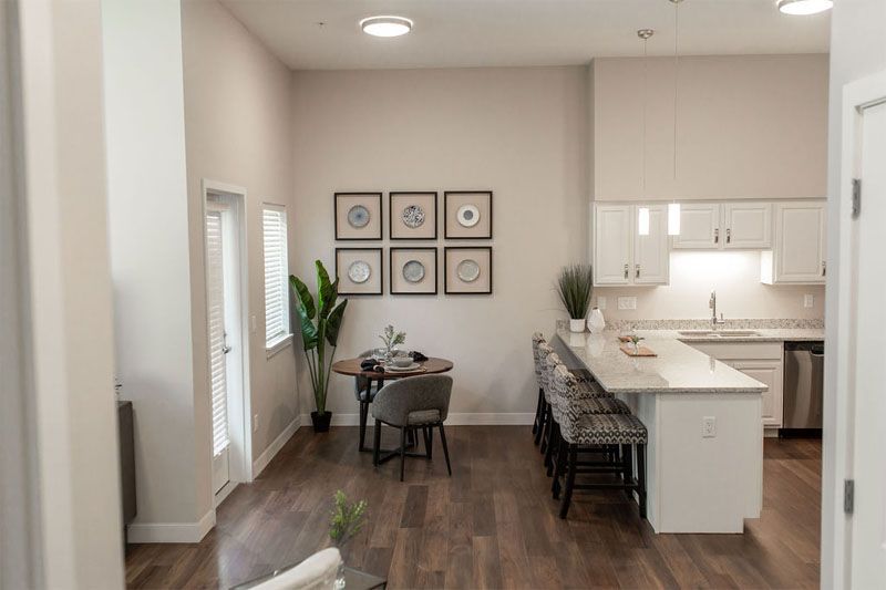 A modern open-concept kitchen and dining area with hardwood floors, bar stools, a small round table, potted plant, and wall art.