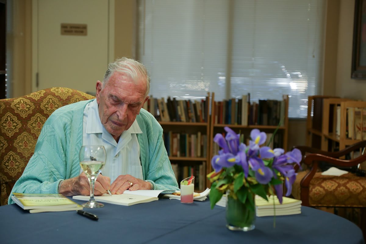 An elderly author signs a book in a library setting with flowers on the table.