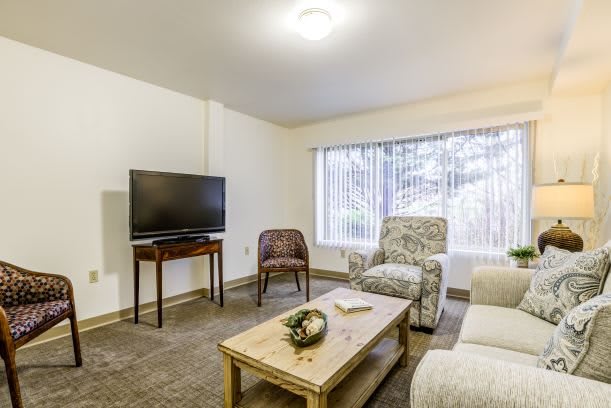 A cozy living room with modern furniture, featuring a television, chairs, a sofa, and a coffee table near a large window.