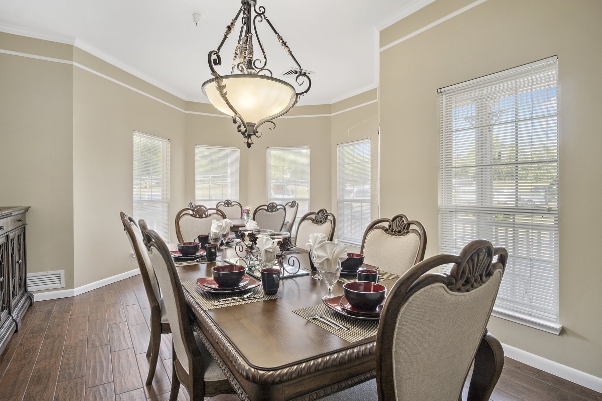 An elegantly furnished dining room with a large table set for a meal, featuring an ornate chandelier and ample natural light from multiple windows.