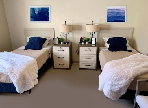 A cozy and minimalist bedroom featuring twin beds with blue pillows, matching nightstands, and calming artwork on the walls.