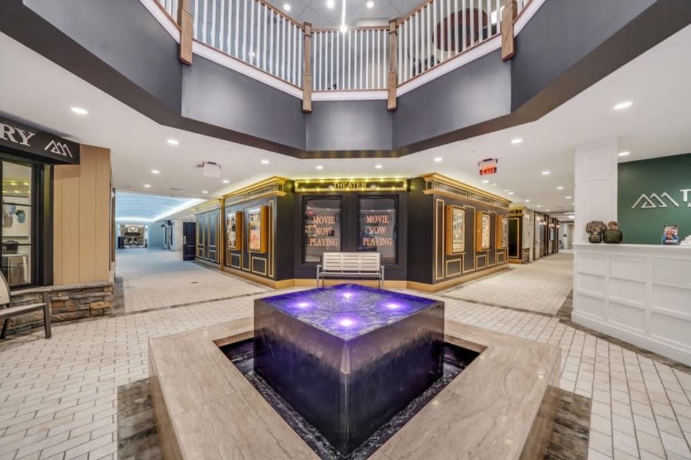 A modern shopping mall interior featuring a central illuminated fountain and elegant retail spaces.