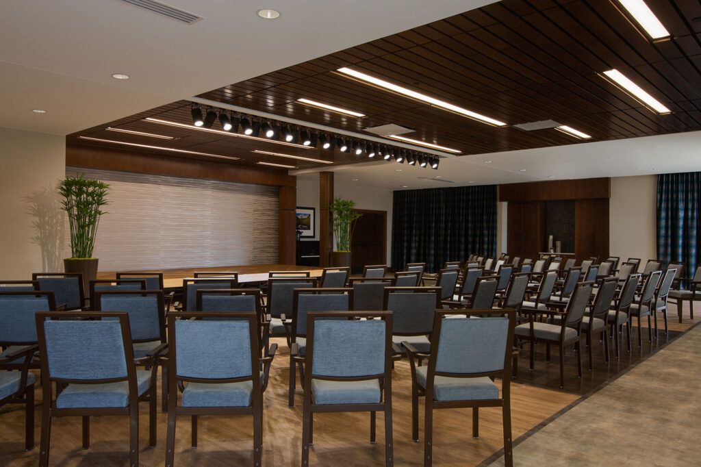 An empty conference room with rows of chairs facing a stage, featuring wooden ceilings and modern lighting.