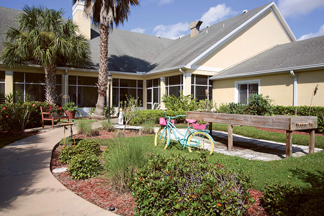 A house with a landscaped garden featuring a decorative bicycle and a palm tree on a sunny day.