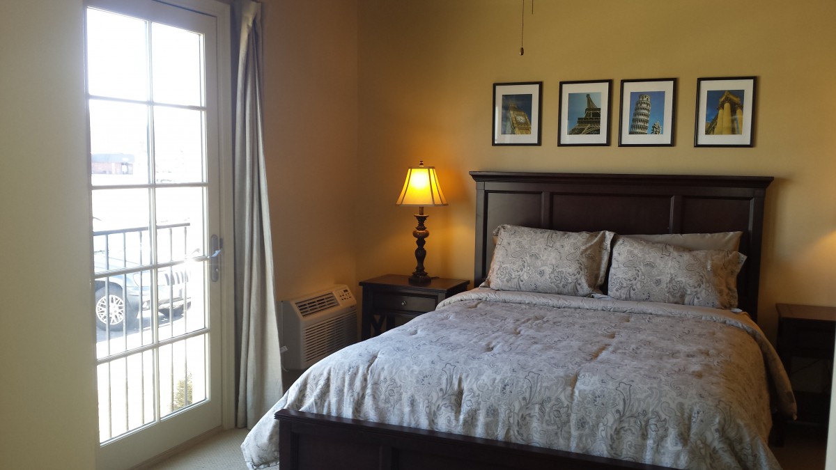 A cozy bedroom with a neatly made bed, nightstand, and lamp beside a window.