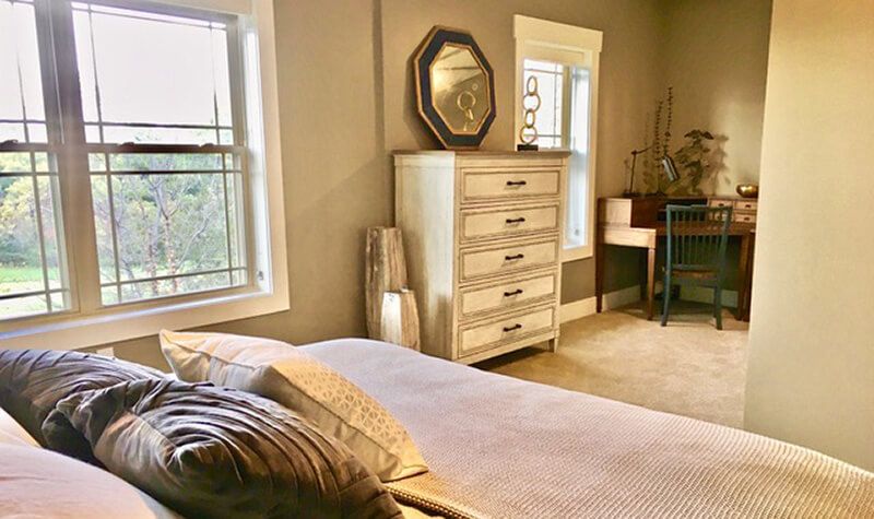 A cozy bedroom with a neutral palette, featuring a bed, a dresser with an octagonal mirror, and two large windows.
