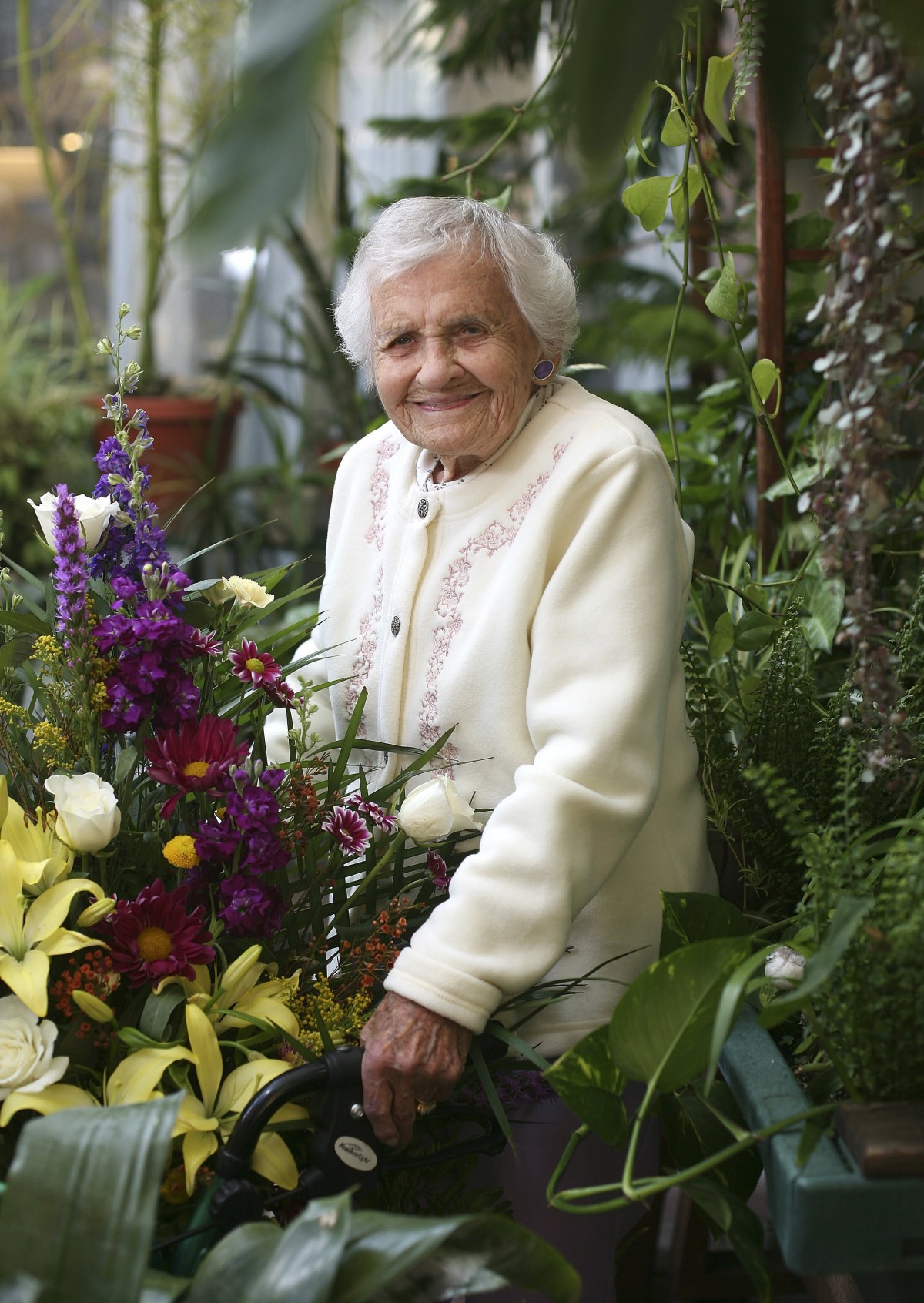 An elderly woman smiling in a lush indoor garden filled with colorful flowers.