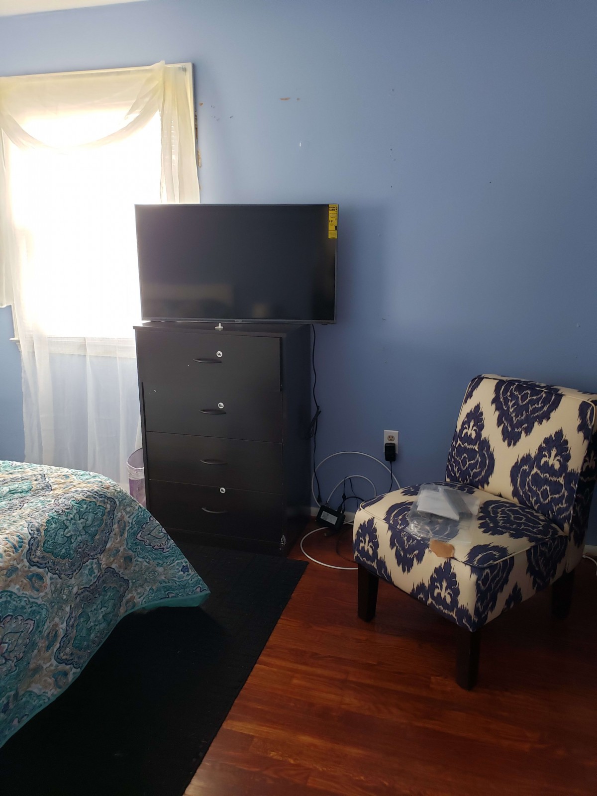 A cozy bedroom with blue walls features a television on a dresser, an armchair, and a bed near the window.