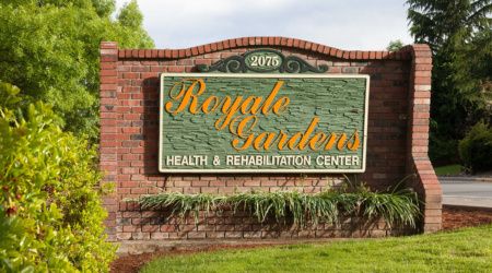 A sign for Royale Gardens Health & Rehabilitation Center, mounted on a brick wall with surrounding greenery.