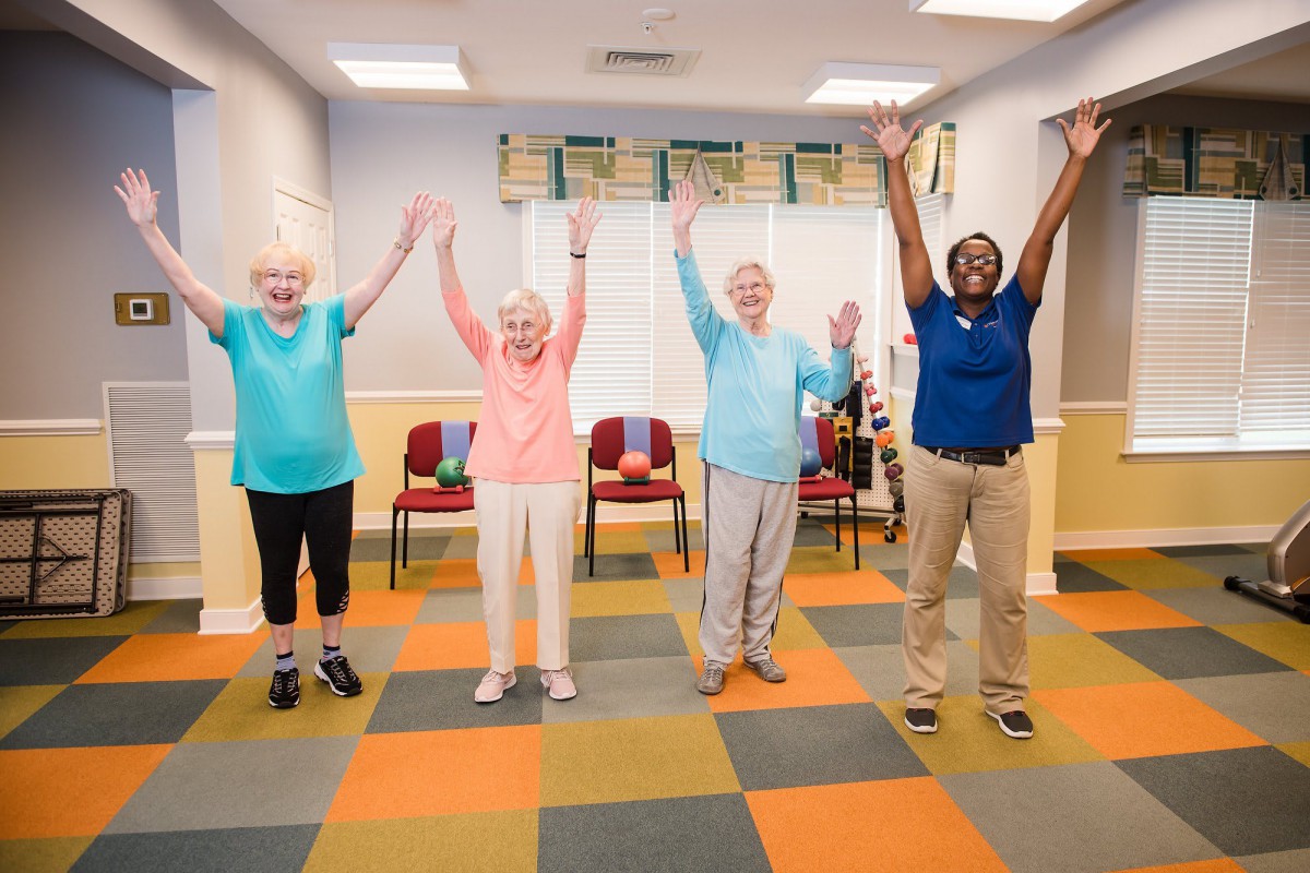 A group of seniors participating in a fitness class indoors, raising their arms while smiling.
