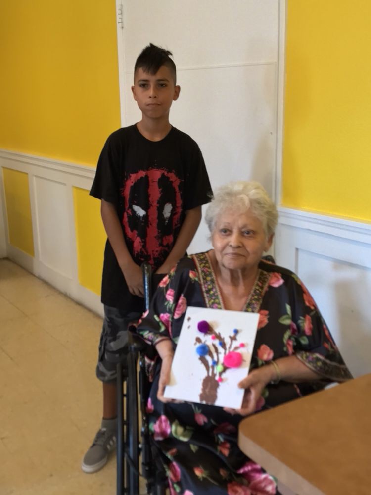 An elderly woman in a wheelchair holds up a colorful piece of artwork, with a young boy standing beside her against a brightly painted yellow wall.