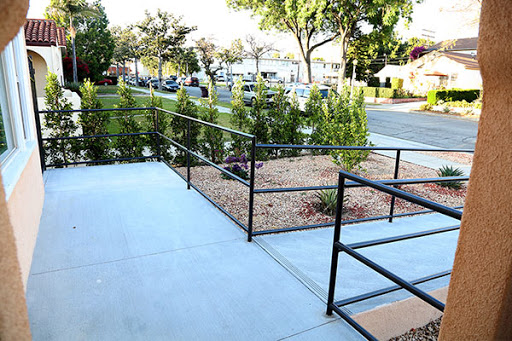 A view of a well-maintained outdoor area featuring a concrete walkway, black handrails, and neatly arranged greenery.