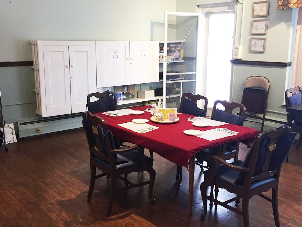 A dining room with a table covered in a red tablecloth, surrounded by chairs and set with placemats. The room features wooden floors and storage cabinets.