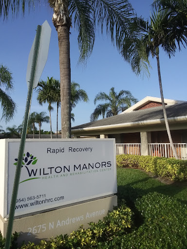 A view of the Wilton Manors facility sign surrounded by palm trees and well-maintained landscaping.