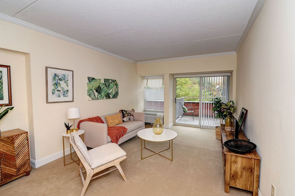 A cozy living room with modern decor, featuring a sofa, coffee table, and artwork on the walls. The space opens up to a balcony with natural light streaming in.
