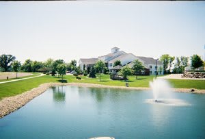A scenic view of a large building near a pond with a fountain, surrounded by lush green landscaping and trees under a clear sky.