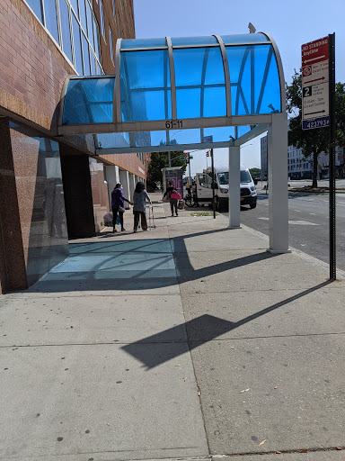 A sunny urban scene featuring a sidewalk with a blue-covered area and a bus stop.