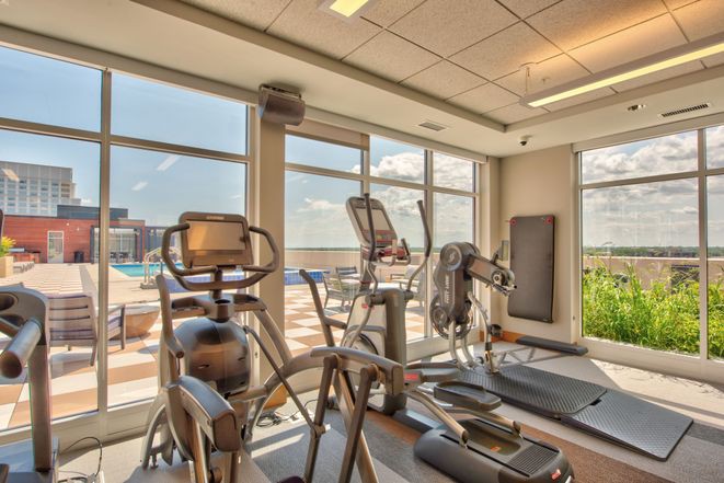 A well-lit gym with exercise equipment including treadmills and ellipticals, overlooking an outdoor view through large windows.