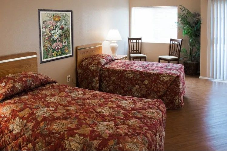 A hotel room with two double beds featuring floral bedspreads, a lamp, and chairs by the window.