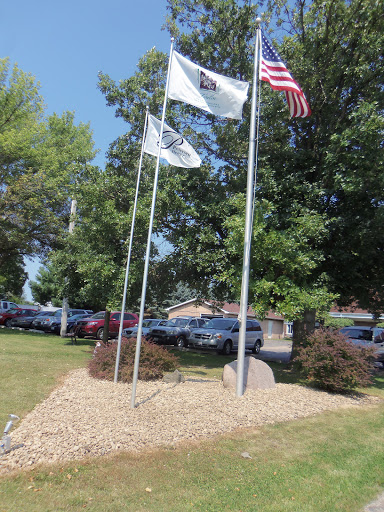 A display of flags in a landscaped area, surrounded by trees and a parking lot.