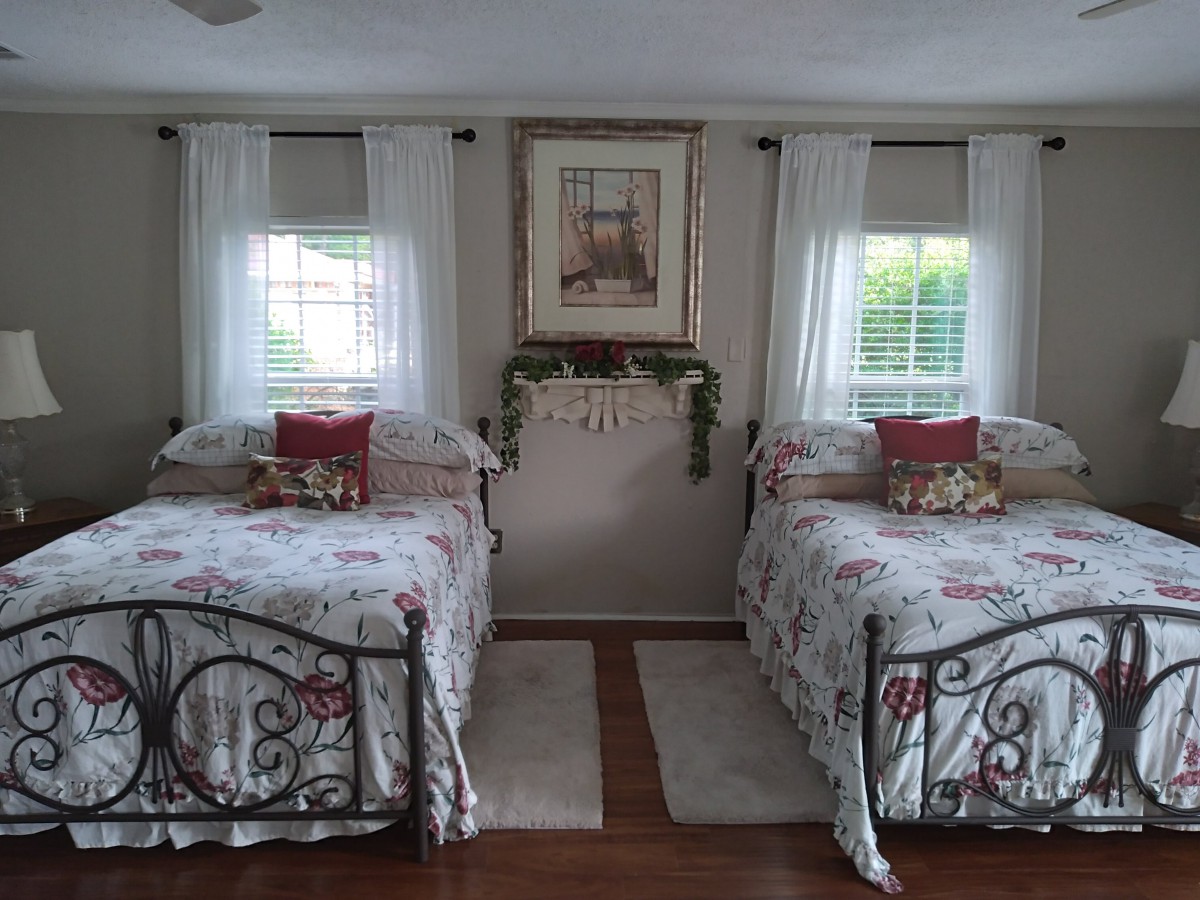 A cozy bedroom with two twin beds, adorned with floral bedding and decorative pillows, flanked by windows with curtains.