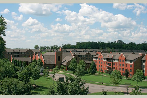 Aerial view of a campus with several buildings surrounded by trees and greenery under a partly cloudy sky.