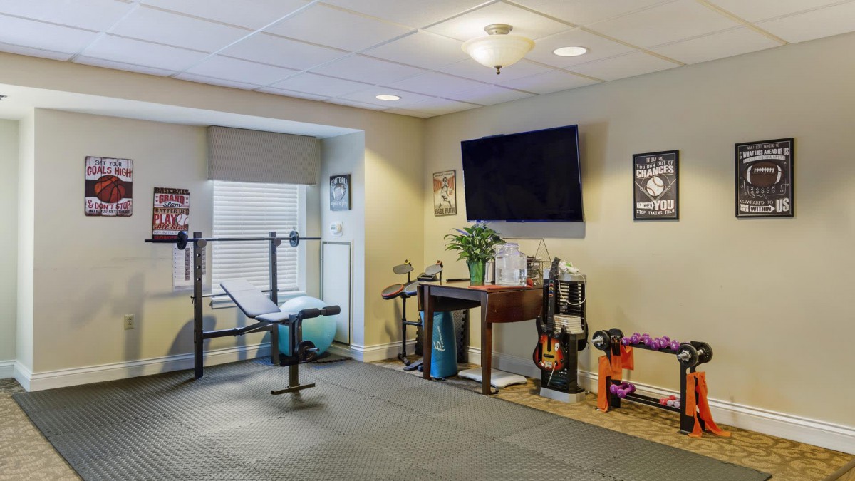 A well-equipped home gym with various exercise equipment, including dumbbells, a barbell rack, and a yoga mat, features a wall-mounted television.