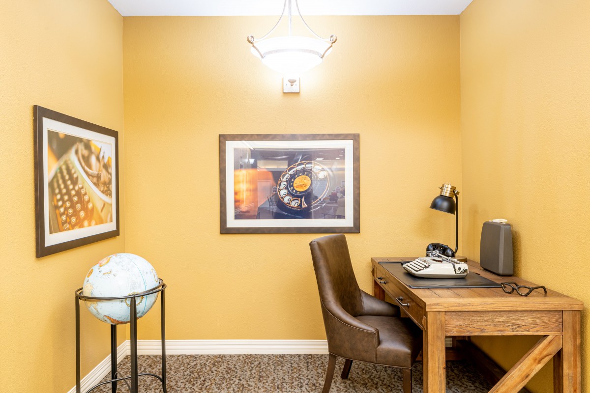 A cozy home office with a wooden desk, chair, lamp, and wall art against yellow walls.