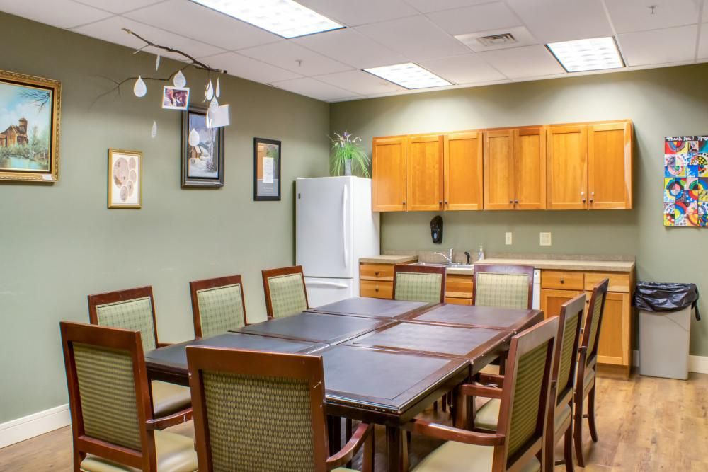 A conference room with a large table surrounded by chairs, adjacent to a kitchen area with cabinets and a refrigerator.