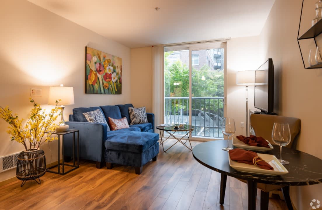 A cozy living room featuring a blue sofa, colorful artwork, and large windows allowing natural light.