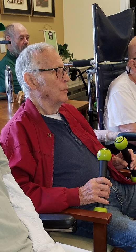 An elderly man wearing glasses and a red jacket, seated in a wheelchair indoors.