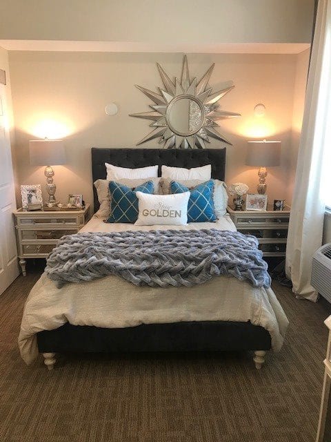A beautifully decorated bedroom featuring a neatly made bed with layered bedding, decorative pillows, and a stylish sunburst clock above the headboard.