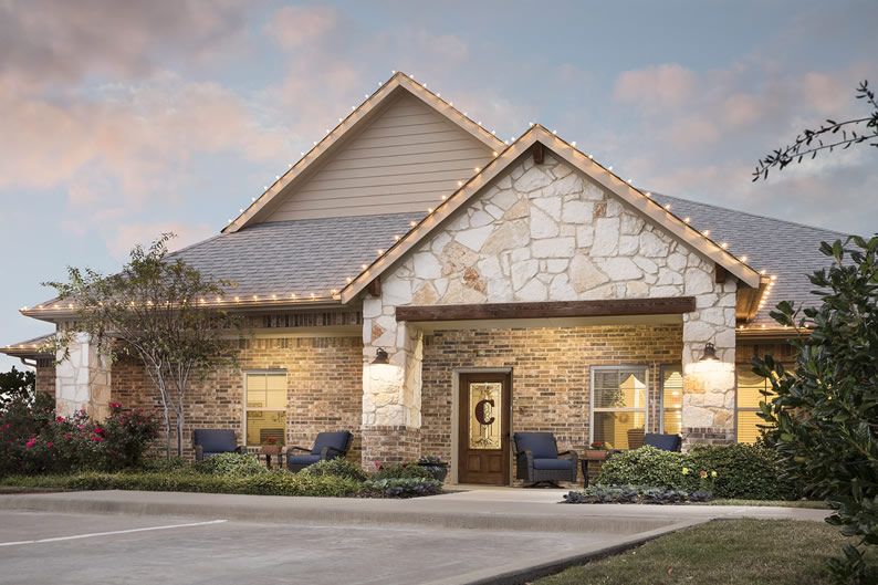 A cozy stone house with a well-lit porch and neatly arranged landscaping, captured during the evening.