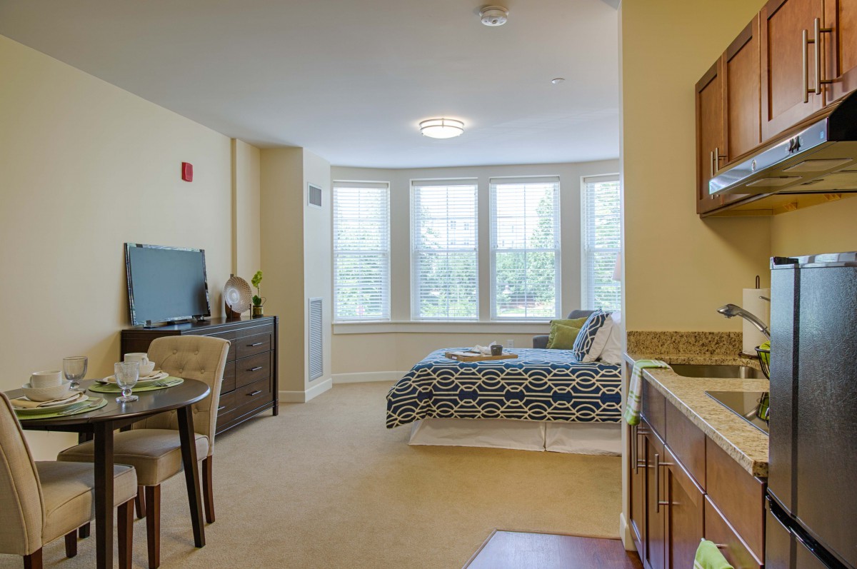 A cozy studio apartment featuring a bed, dining area, and large windows providing ample natural light.