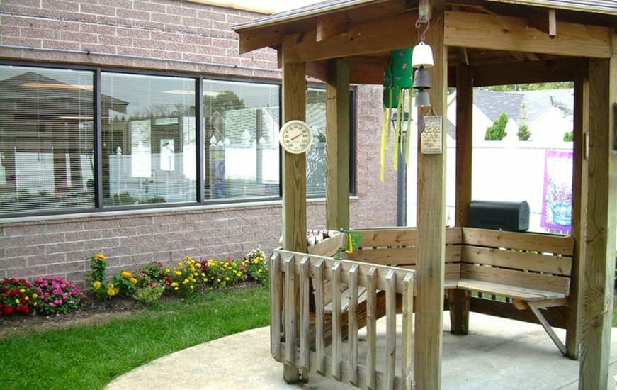 A small wooden gazebo with wind chimes is situated near a brick building, surrounded by a neatly maintained garden.