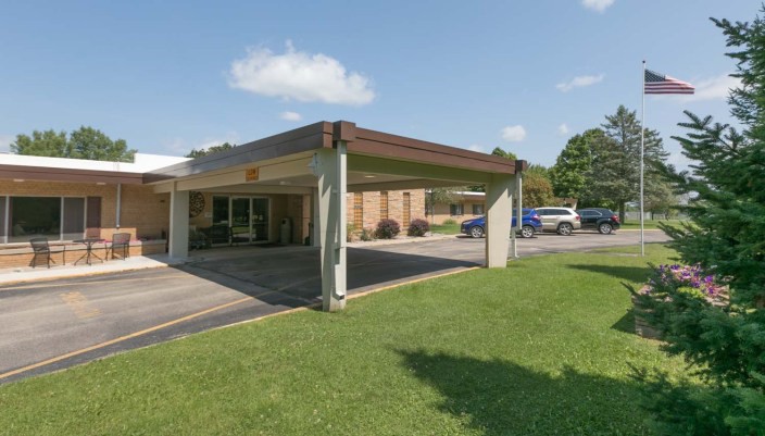 A building with a covered entrance and adjacent parking lot, surrounded by grass and trees under a partially cloudy sky.
