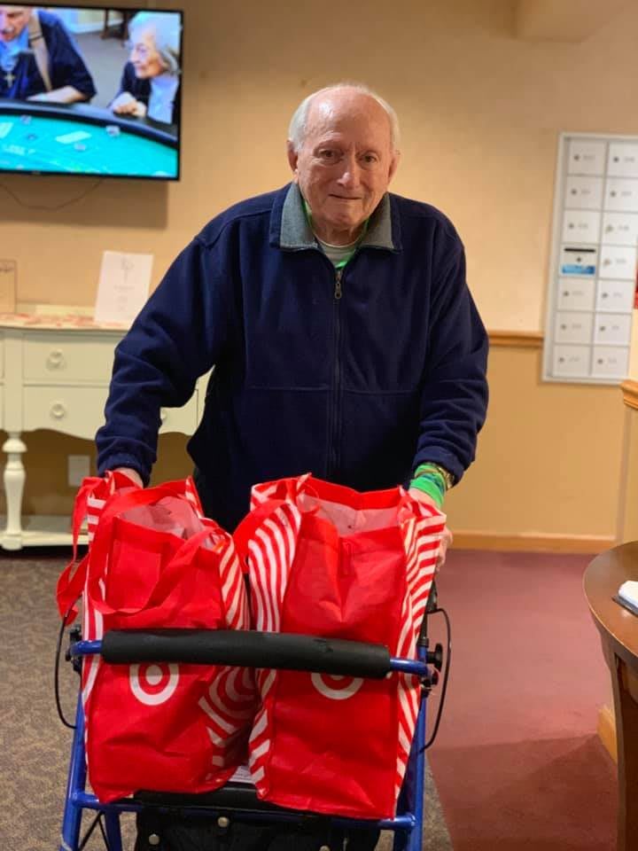 An elderly person using a walker carries shopping bags indoors, smiling.