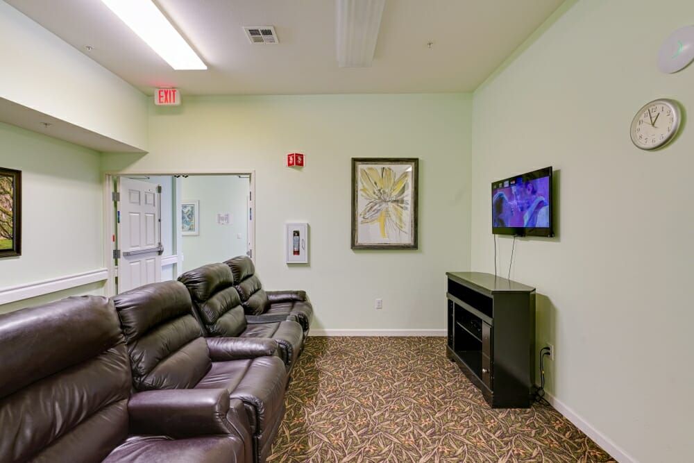 A cozy living room with recliner chairs facing a mounted television above a fireplace, featuring a carpeted floor and framed artwork on the wall.