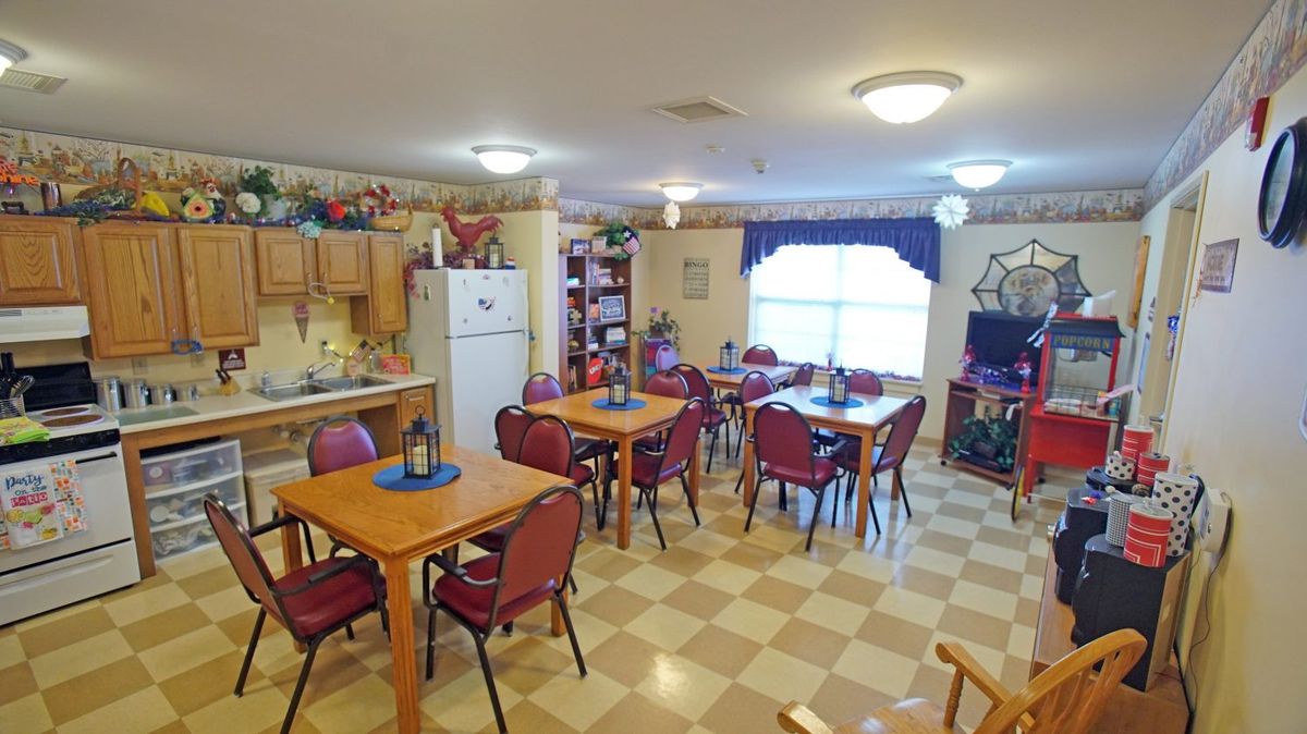 A cozy dining room with multiple tables and chairs, featuring a small kitchen area and warm decorations.