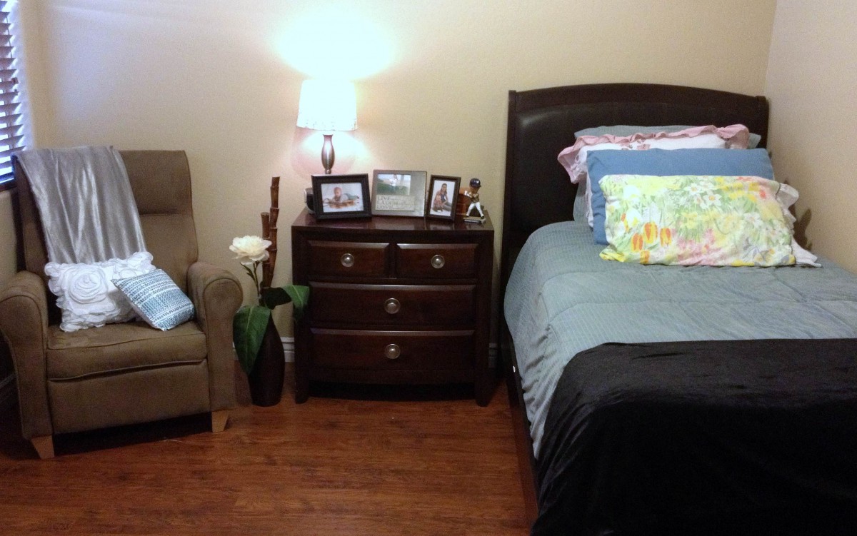A cozy bedroom with a neatly made bed, nightstand with photo frames and a lamp, and an armchair with decorative pillows.