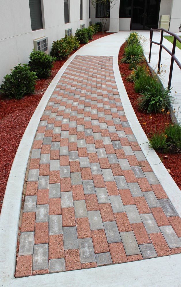 A neatly laid brick pathway with a geometric pattern, bordered by landscaped plants and red mulch.