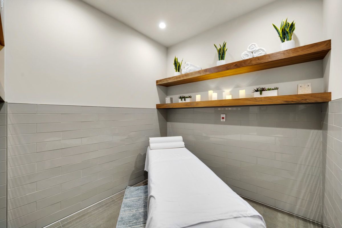 A serene spa room featuring a massage table and minimalist decor with plants on shelves.