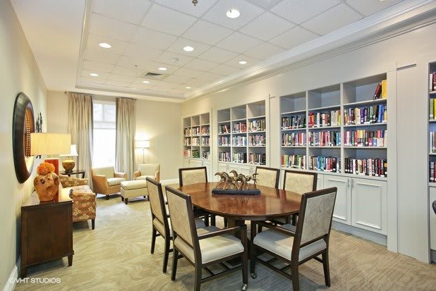 A cozy library room featuring a round table surrounded by chairs, with bookshelves lining the walls and warm lighting creating an inviting atmosphere.