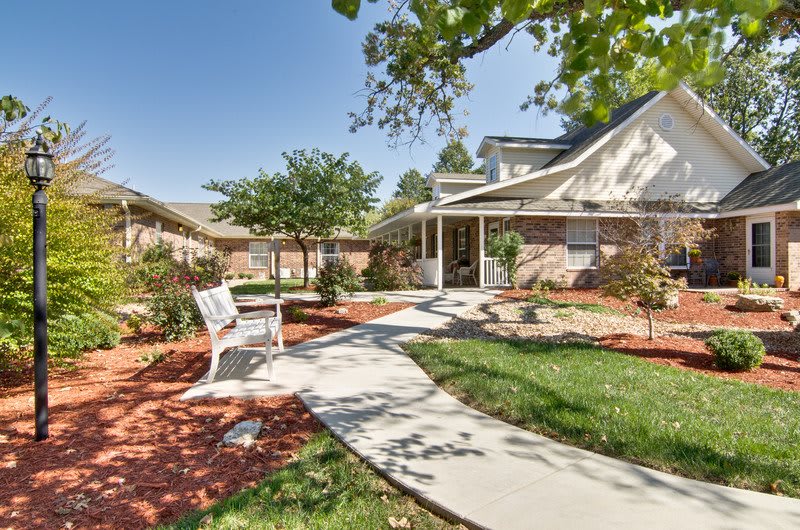 A well-maintained garden with a paved pathway leading to a house, featuring trees and a bench.