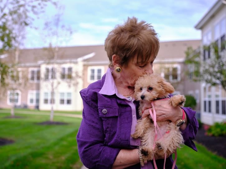 An elderly woman affectionately holding a small puppy outdoors in front of a building.