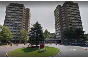 Two tall residential apartment buildings with a central entrance sign and trees in the foreground.