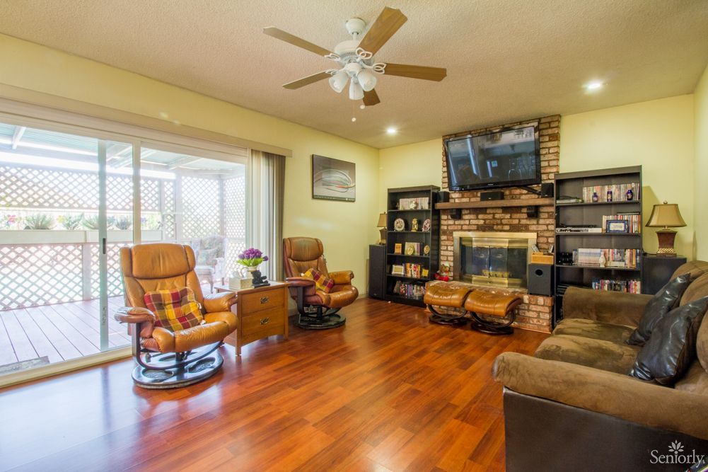 A cozy living room with wooden flooring, featuring a fireplace, television, armchairs, and a ceiling fan.