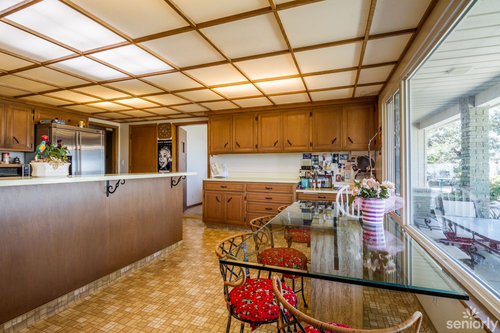 A retro-style kitchen with wooden cabinets, a glass table, and red chairs, featuring a vintage 1970s design.