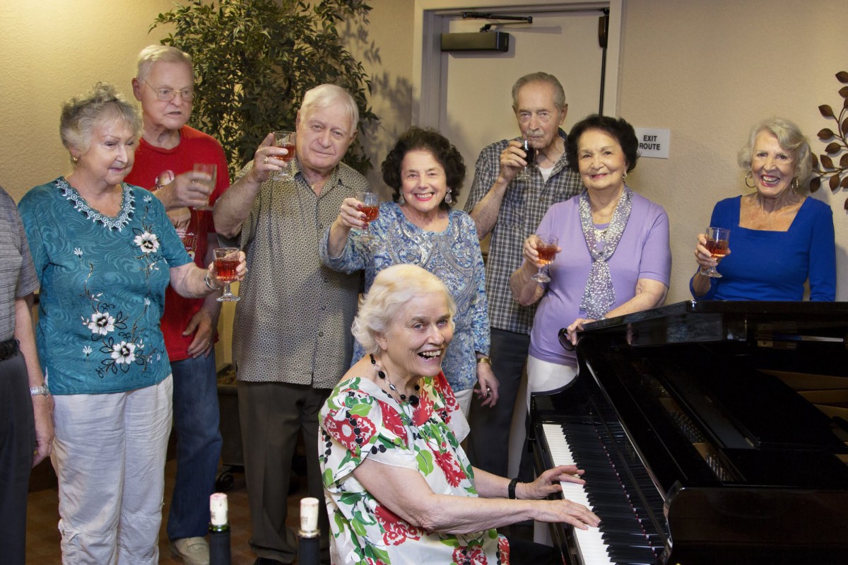 A group of elderly people celebrating with drinks while a woman plays the piano.